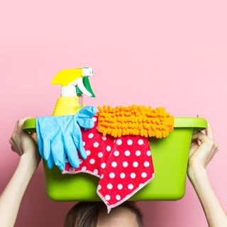 A person - who is not seen besides their forehead and hands - holds a green plastic tub on top of their head with cleaning supplies including two spray bottles, gloves, and some colorful rags.