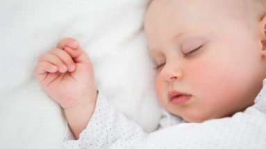 An up close image of a baby sleeping on its back with hands balled up above its head.