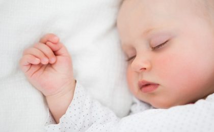 An up close image of a baby sleeping on its back with hands balled up above its head.
