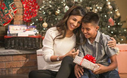 A woman and boy - presumably mother and son - sit together under a Christmas tree. The boy is holding a wrapped present. They are both smiling, the woman's arm is around the boy's shoulder.
