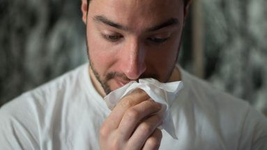A close up image of a man on the verge of blowing his nose, he is holding a tissue up to his face and facing down.