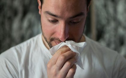 A close up image of a man on the verge of blowing his nose, he is holding a tissue up to his face and facing down.