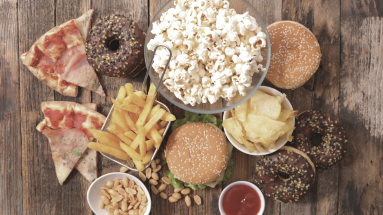 An aerial view of a wooden table with a bunch of junk food on it, including French fries, popcorn, pizza, donuts, peanuts, potato chips, and hamburgers.