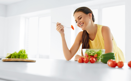 A woman in a white kitchen sits at the counter with a bowl of salad.