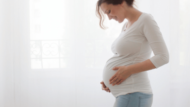 A pregnant white woman in a white shirt and blue jeans gazes down at her belly.