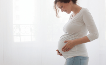 A pregnant white woman in a white shirt and blue jeans gazes down at her belly.