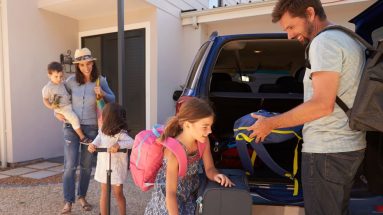 A smiling young family with mother, father, two daughters, and a son pack up an SUV for a road trip with suitcase and bags.