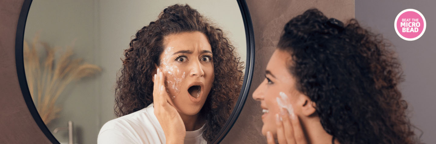 The image has two pictures of the same woman in a bathroom– one is in a mirror. On the right side, we can see her profile. She is smiling and applying white lotion. On the left side, in the mirror, we can see the same woman from the front. Instead of a smile, she has a shocked look on her face with her mouth gaping open. There’s a layer of clear plastic on her cheek where the mirror image has lotion. “Beat the Microbead” is in the top right corner in a pink circle with white text.