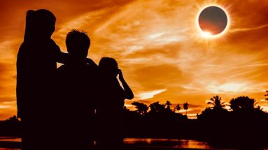 A family in the foreground takes in a total solar eclipse in the background of a tropical panorama bathed in orange light.
