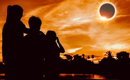 A family in the foreground takes in a total solar eclipse in the background of a tropical panorama bathed in orange light.