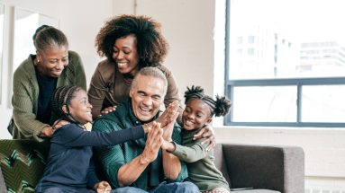 Members of three generations of an African-American family are together. A well-dressed elderly man sitting on a couch high fives with two girl children, one sitting on either side of him, as two women standing behind the couch watch. One of the women is also elderly.