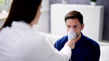 A white man with blue hair in a royal blue button down shirt sits in a health clinic and receives treatment from a nebulizer from a woman doctor whose back is to the viewer.