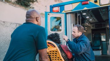 Two male paramedics attend to a person on a stretcher in front of open ambulance doors, prior to putting the person inside the back. It is a sunny day.