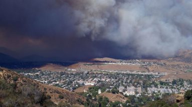 A daytime scene of a valley town close to a wildfire. There is a huge cloud of smoke over the town and to the left the sky is very dark.