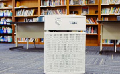 A close up of a sandstone Austin Air purifier in an elementary school library, there are bookshelves full of books and two desks in the background. In the back left there is a bookshelf that has colorful bins with magazines and soft cover books.
