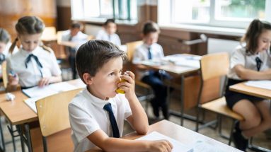 A scene in an elementary classroom featuring a young boy with brown hair breathing with an inhaler. The boy and his classmates are wearing school uniforms of black pants/skirts, white shirts, with ties.