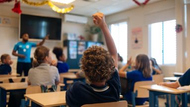 A photo taken from the back of an elementary school classroom shows a Black man teacher with children in discussion, one child at the back of the room has his hands raised.