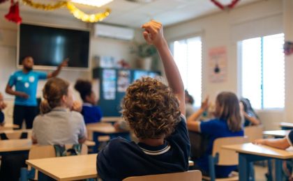 A photo taken from the back of an elementary school classroom shows a Black man teacher with children in discussion, one child at the back of the room has his hands raised.