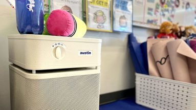 A close up image of a sandstone-colored Austin Air purifier inside an elementary school classroom, with toys and teaching supplies in the background.