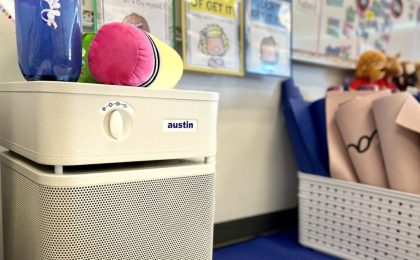 A close up image of a sandstone-colored Austin Air purifier inside an elementary school classroom, with toys and teaching supplies in the background.