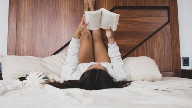 A woman with brown hair in comfy clothes is in an unmade bed with white sheets. Her legs up on the wooden headboard and she is lost in a book.