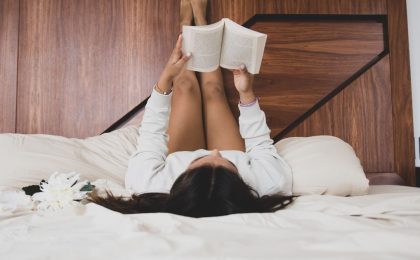 A woman with brown hair in comfy clothes is in an unmade bed with white sheets. Her legs up on the wooden headboard and she is lost in a book.