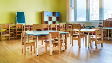 A photo of a classroom for very small children with small desks and cubby spaces. The room is lit up by natural light from a window on the right and there is a white skyscraper outside. There are toy trucks visible to the right, up against the wall.