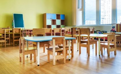 A photo of a classroom for very small children with small desks and cubby spaces. The room is lit up by natural light from a window on the right and there is a white skyscraper outside. There are toy trucks visible to the right, up against the wall.