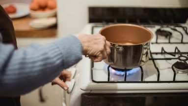 Close up of a white gas stove, showing a person wearing a blue sweater placing a saucepan with water in it on the burner with their right hand and turning the stove knob with their left hand.