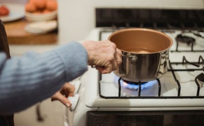 Close up of a white gas stove, showing a person wearing a blue sweater placing a saucepan with water in it on the burner with their right hand and turning the stove knob with their left hand.