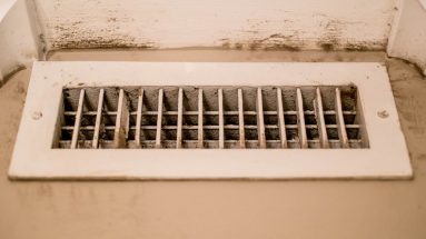 A close up of a dirty heating/cooling vent. It is on the floor and the vent itself is dusty, the area around it also shows a lot of built up dirt on the white trim around the vent.