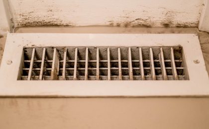 A close up of a dirty heating/cooling vent. It is on the floor and the vent itself is dusty, the area around it also shows a lot of built up dirt on the white trim around the vent.