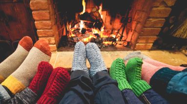 A photo of a wood burning fireplace with a roaring fire. A family is snuggled up in front of the fireplace with three small pairs of feet (kids) and two larger pairs of feet (adults) warming by the fire, wearing thick colorful socks. (Only their legs and feet are visible.)