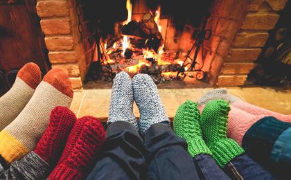 A photo of a wood burning fireplace with a roaring fire. A family is snuggled up in front of the fireplace with three small pairs of feet (kids) and two larger pairs of feet (adults) warming by the fire, wearing thick colorful socks. (Only their legs and feet are visible.)