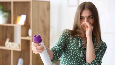 A brunette woman in a green polka dotted dress covers her nose and makes a disgusted face while reading a can of aerosol air freshener. She is in a white room with some light wood shelving in the back left.