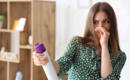 A brunette woman in a green polka dotted dress covers her nose and makes a disgusted face while reading a can of aerosol air freshener. She is in a white room with some light wood shelving in the back left.