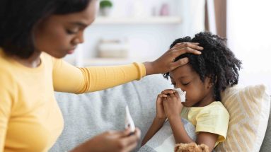 A Black mother tends to her sick daughter who is lying on a couch. The mother appears concerned as she looks at a thermometer and holds her other hand to her child’s forehead. The little girl has a tissue held up to her nose, she’s under a blanket, reclining on a yellow pillow, and there is a teddy bear by her side with a blue bow around its neck.