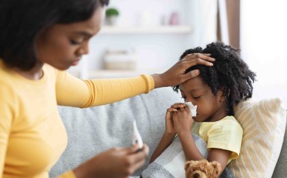 A Black mother tends to her sick daughter who is lying on a couch. The mother appears concerned as she looks at a thermometer and holds her other hand to her child’s forehead. The little girl has a tissue held up to her nose, she’s under a blanket, reclining on a yellow pillow, and there is a teddy bear by her side with a blue bow around its neck.