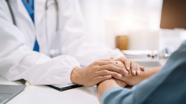 A medical setting, only two sets of hands are visible. A doctor in a white coat is on the left of the image, facing the camera, reaching out to hold the hands of a patient who has their back to the camera.