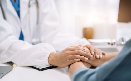 A medical setting, only two sets of hands are visible. A doctor in a white coat is on the left of the image, facing the camera, reaching out to hold the hands of a patient who has their back to the camera.