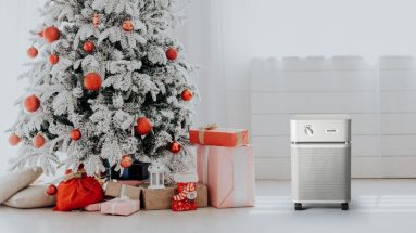 A beautiful Christmas tree with well wrapped colorful presents beneath it. The tree is in a bright white room and looks snow covered but is indoors. The ornaments and presents are all in a similar coral and light pink color scheme. There is an Austin Air Purifier in sandstone to the right of the Christmas tree.