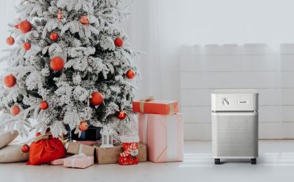 A beautiful Christmas tree with well wrapped colorful presents beneath it. The tree is in a bright white room and looks snow covered but is indoors. The ornaments and presents are all in a similar coral and light pink color scheme. There is an Austin Air Purifier in sandstone to the right of the Christmas tree.