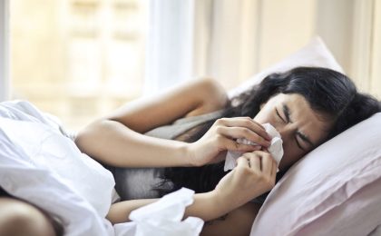 A woman with long brown hair lays on her side in bed. She is blowing hard into a tissue, her eyes are closed. She appears to be sick. The bed lines are white. She is wearing a gray wide strap tank top.