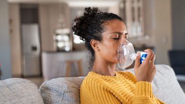 A woman uses a nebulizer for a respiratory treatment. She is a woman of color and has curly hair that is in a bun. She is wear a yellow sweater and sitting on a light gray couch. You can see a kitchen in the background.