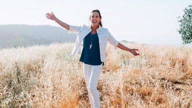 A beautiful brunette woman stands in a golden field on a bright sunny day. She is Dr. Jill Carnahan and she is wearing white jeans and a white jacket with a jewel blue shirt. She is smiling and has her hands out. There is a tree in the background on the right.