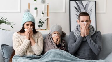 A family of three sits on a couch, with a girl around the age of 10 sitting in the center of her parents. They are sharing a blanket, the mother and daughter are each wearing knit hats. The father is wearing a large knit scarf. All three of them are blowing their noses. They are in a white room with some houseplants in the background.