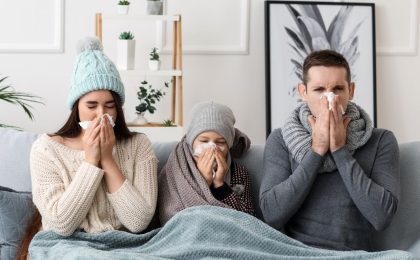 A family of three sits on a couch, with a girl around the age of 10 sitting in the center of her parents. They are sharing a blanket, the mother and daughter are each wearing knit hats. The father is wearing a large knit scarf. All three of them are blowing their noses. They are in a white room with some houseplants in the background.