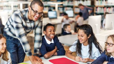 A smiling group of schoolchildren is sitting in a library with a friendly-looking young man instructor bending down to be closer to them. The kids are in basic uniforms consisting of white polo shirts. One boy and one girl are also wearing navy blue long sleeve shirts. There are more kids and another adult in the background, to the right, but they are blurry.