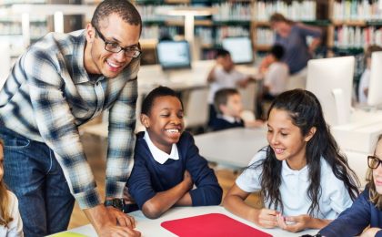 A smiling group of schoolchildren is sitting in a library with a friendly-looking young man instructor bending down to be closer to them. The kids are in basic uniforms consisting of white polo shirts. One boy and one girl are also wearing navy blue long sleeve shirts. There are more kids and another adult in the background, to the right, but they are blurry.