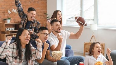 A group of people is watching a football game on TV and cheering whole eating popcorn and potato chips. We can guess they are watching football because one of the viewers is holding a football. It is a bright day, the sun is showing through the windows.